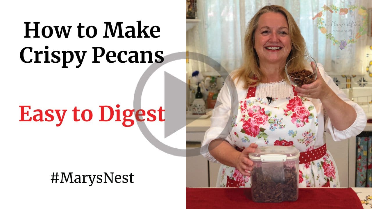 Mary in her kitchen behind a counter with a container of crispy nuts and she's holding some in a cup.